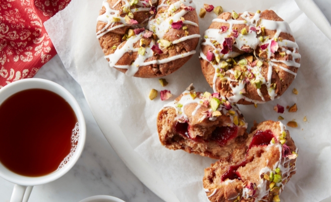 Raspberry rose scone-muffins garnished with vanilla icing drizzle, dried rose pedals, and chopped pistachios. All on a white platter with a side cup of tea and bowl of pistachios.