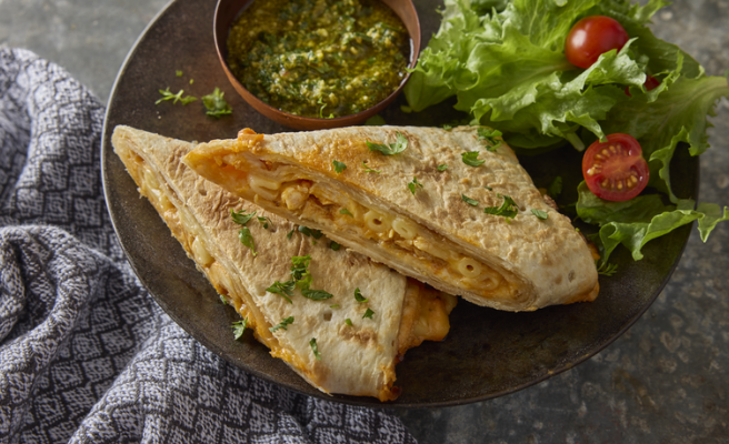 Two mac n’ cheese wraps stacked on a plate with a small side salad and small bowl of cilantro chutney sauce.