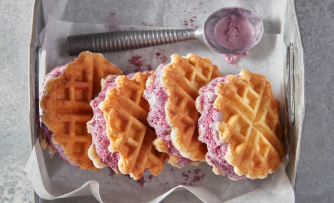 Four waffled biscuit ice cream sandwiches on parchment paper and silver serving tray with ice cream scoop.