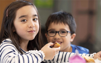 Two kids smile at the camera while eating a meal together.