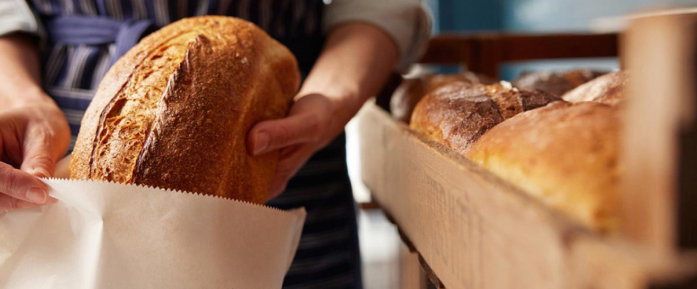 A person places a loaf of bread into a paper bag beside a wooden tray filled with freshly baked bread.